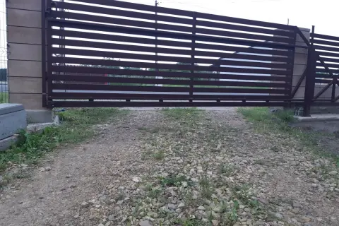 Brązowa, metalowa brama przesuwna z poziomymi szczeblinami. Widoczna jest szczelina pod bramą, przez którą widać trawę i żwir.