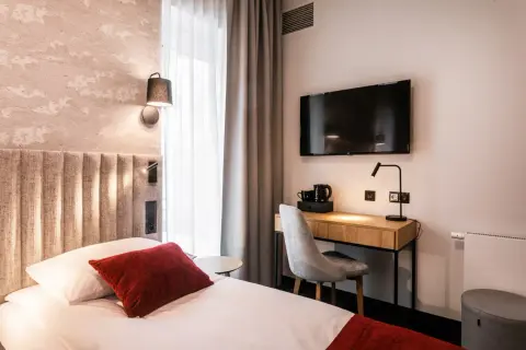 Przytulny pokój w hotelu Vesper House: łóżko z czerwonymi akcentami, biurko z czajnikiem i telewizorem, szare zasłony.