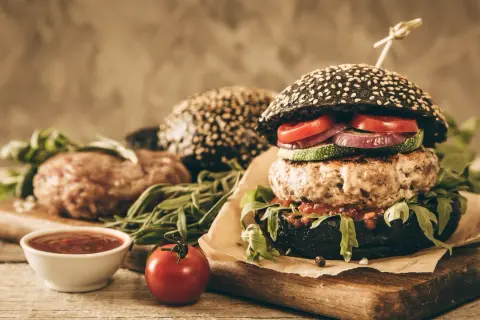 Gotowy hamburger z czarną bułką, kotletem wołowym i warzywami, obok surowy kotlet i sos.
