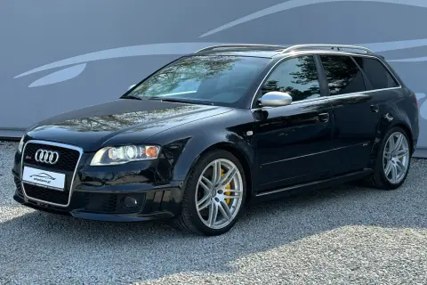 Czarny Audi RS4 B7 Avant z żółtymi zaciskami hamulcowymi, zaparkowany na żwirze przed szarą ścianą z grafiką.