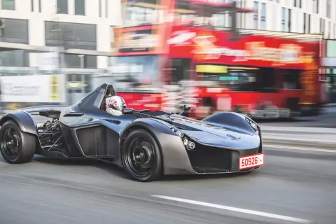 Szary BAC Mono z kierowcą w kasku pędzi ulicą, mijając rozmazany czerwony autobus piętrowy.