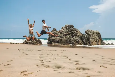 Szczęśliwa para skacze na plaży w meksykańskim kurorcie nad Pacyfikiem, z falami i skałami w tle.