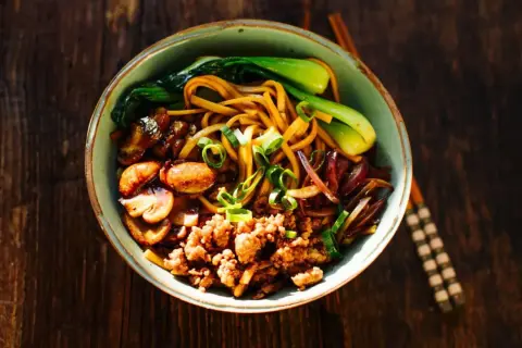 Herzhafte Udon Nudeln mit gebratenem Tofu, Pilzen, Pak Choi und Frühlingszwiebeln auf einem rustikalen Holztisch.