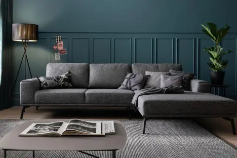 Ein modernes Wohnzimmer mit einem grauen Sofa, das mit Kissen und einer Decke dekoriert ist. Ein Stehlampen-Design und eine Zimmerpflanze runden das Bild ab.