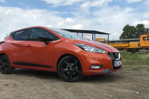 Pomarańczowy Nissan Micra na tle torów kolejowych. Sprawdź nissan micra wymiary, by poznać jego kompaktowe gabaryty.