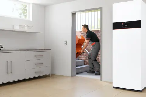 Instalator wnosi nowy, pomarańczowy element, który może być częścią odpowiedzi na pytanie: jaki kocioł gazowy do domu? Obok stoi nowoczesny, biały kocioł.