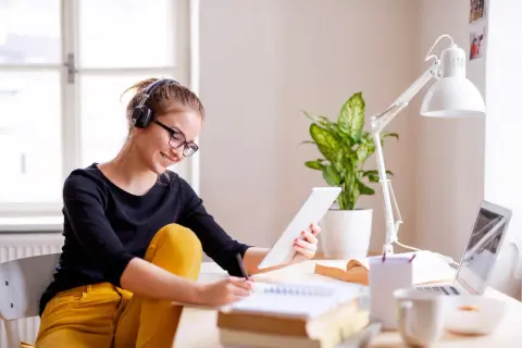 Junge Frau mit Kopfhörern und Brille lernt am Tablet für ihr **vollzeit studium**. Sie schreibt Notizen in ein Heft.