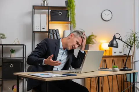 Zestresowany mężczyzna w garniturze trzyma się za głowę, patrząc na laptopa. Może właśnie stracił wadium.