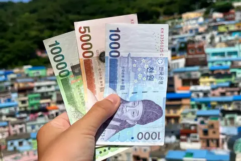 Dłoń trzyma banknoty wonów, pokazując korea południowa ceny. W tle kolorowe domy na wzgórzu.