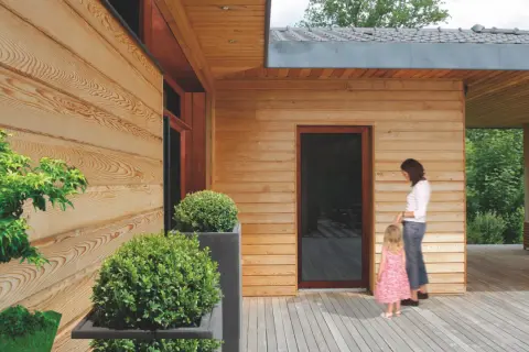 Drewniana elewacja domu, zabezpieczona impregnatem, z kobietą i dzieckiem na tarasie.