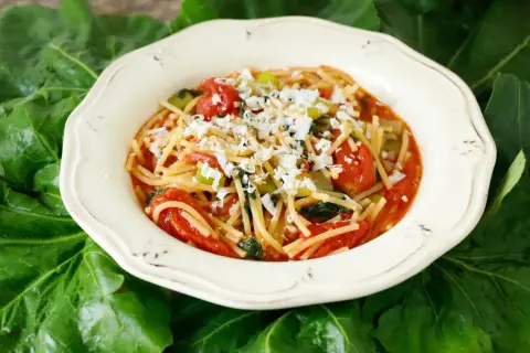 Un piatto di pasta con i tenerumi asciutta, condita con pomodoro fresco e formaggio grattugiato, adagiato su foglie verdi.