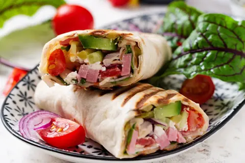 Pyszna tortilla z szynką, awokado, pomidorami i serem feta, podana z sałatką i czerwoną cebulą.