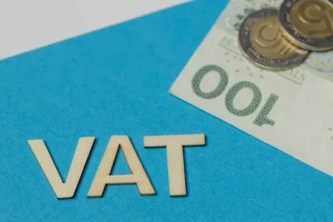 Drewniane litery VAT na niebieskim tle, obok banknot 100 zł i monety. Rozważanie vat należny a naliczony.