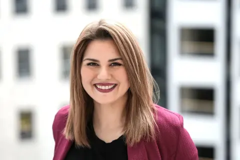 Lächelnde junge Frau mit braunen Haaren, die einen pinken Blazer trägt. Sie studiert IU Master Wirtschaftsinformatik und blickt optimistisch in die Zukunft.
