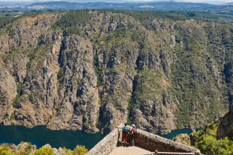 Dos excursionistas contemplan la majestuosidad de los ríos de Galicia, serpenteando entre cañones rocosos y bosques frondosos.