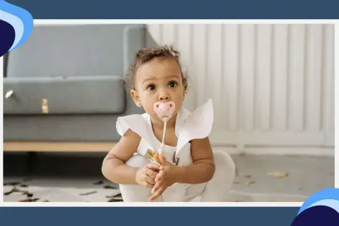 A baby girl with a pink pacifier in her mouth sits on the floor, her eyes wide with curiosity. The pacifier terth is a comforting presence as she explores her surroundings.