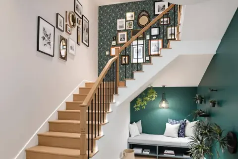 Un habillage mural de cage d'escalier avec une galerie de cadres et un papier peint géométrique vert. Un banc confortable est aménagé sous l'escalier.