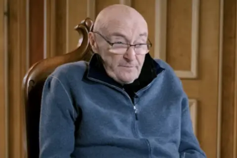 Phil Collins, zmagający się z chorobą, siedzi w fotelu, ubrany w niebieski polar.