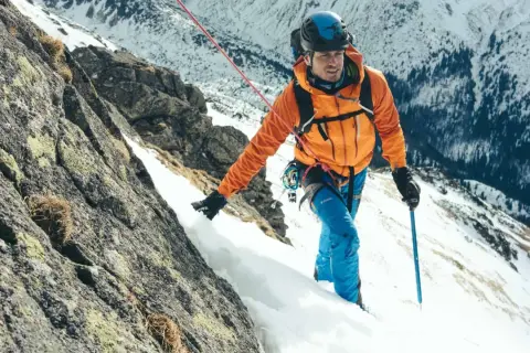 Alpinista w pomarańczowej kurtce wspina się po skalistym, ośnieżonym zboczu, używając czekana alpinisty inaczej jako wsparcia.