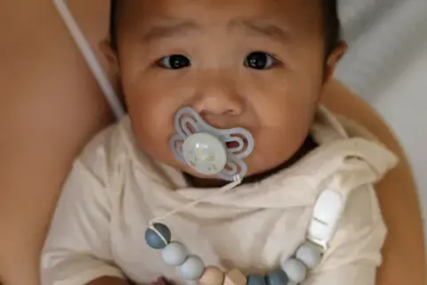 A baby with a pacifier, possibly experiencing pacifier nipple confusion, looks into the camera while being held by a parent.