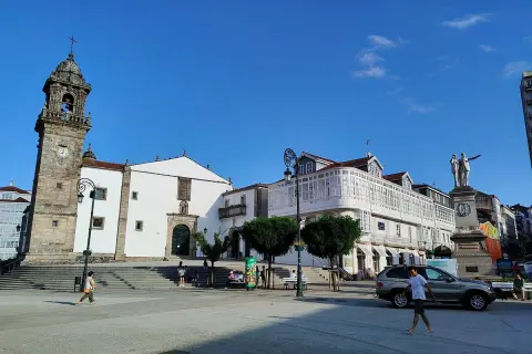 La plaza Betanzos, con su iglesia de torre campanario, edificios blancos y un monumento, bajo un cielo azul.
