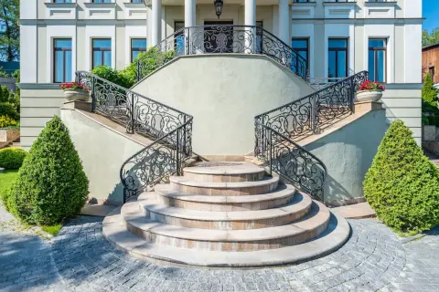 Imponente villa con scale esterne coperte da ringhiere in ferro battuto e ornate da vasi con fiori rossi.