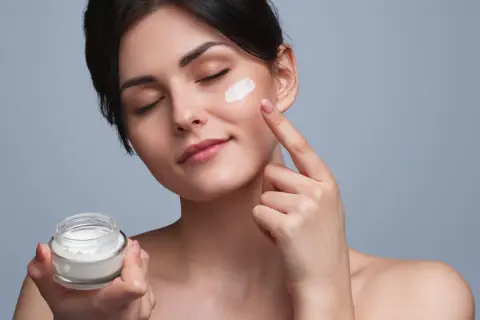 Frau trägt eine Tages- und Nachtcreme auf. Sie hält ein Glas mit der Creme in der Hand und tupft sie mit dem Finger auf ihre Wange.