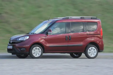 Czerwone kombi auto w ruchu, z widocznymi drzwiami przesuwnymi i relingami dachowymi, na tle rozmytego zielonego krajobrazu.
