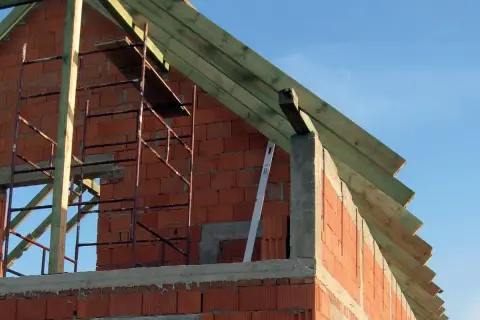 Budowa domu, widok na ceglane ściany i drewniany dach. Widać rusztowanie i elementy konstrukcyjne. Odległość komina od krokwi jest kluczowa dla bezpieczeństwa.
