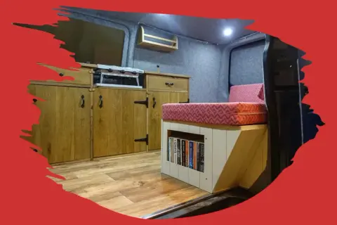 Interior de caravana con cocina, cama y estanterías. El suelo de madera renovado da un aspecto acogedor.