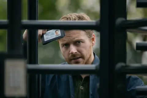 Ein Mann mit blondem Haar und Bart hält einen Ausweis hoch, während er durch ein Gitter blickt. Spannung in Slow Horses Staffel 3.