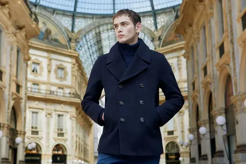 Giovane uomo con un caban blu scuro, un capo che incarna eleganza e calore, passeggia sotto la volta di una galleria storica.