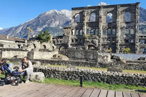 Un uomo con un passeggino ammira le rovine romane, uno dei **paesi più belli della Valle d'Aosta**, con montagne innevate sullo sfondo.