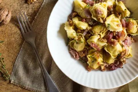 En skål med tortellini prosciutto, valnötter och timjan. En gaffel ligger bredvid på en rustik träyta.