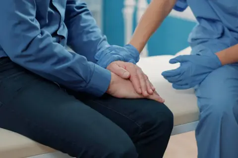 Pielęgniarka w niebieskich rękawiczkach pociesza pacjenta po operacji prostaty, który zmaga się z nietrzymaniem moczu.