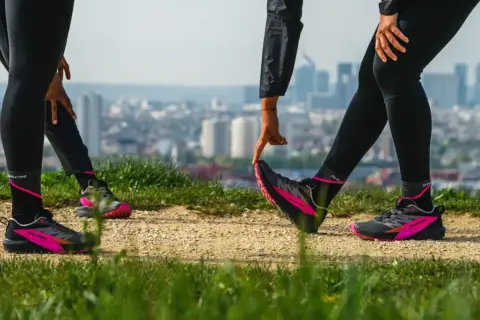 Zwei Läuferinnen dehnen sich auf einem Hügel mit Blick auf eine Stadt. Sie tragen schwarze Laufhosen und auffällige pink-schwarze Laufschuhe. Ein Zeichen für den Wiedereinstieg ins joggen nach Geburt.
