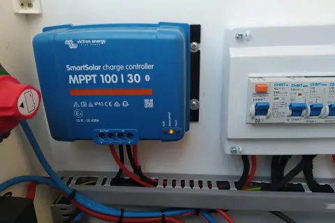 Controlador de carga solar Victron MPPT 100|30, con luces indicadoras de estado y cables conectados. A su derecha, un cuadro eléctrico con interruptores.