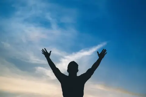 Sylwetka człowieka z rękami w górze na tle błękitnego nieba z chmurami. Jak w psalm 113, uwielbienie.
