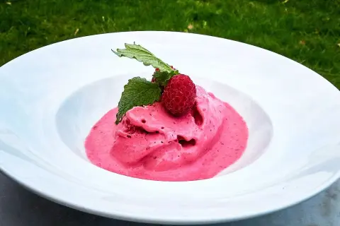 Hallonglass, gjord i en glassmaskin, serveras i en vit skål med färska hallon och mynta.
