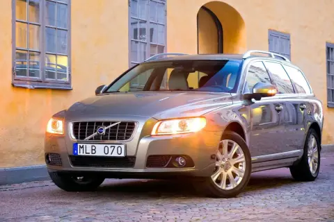 En servad bil, en silverfärgad Volvo kombi, står parkerad på en kullerstensgata framför en gul byggnad med fönster.