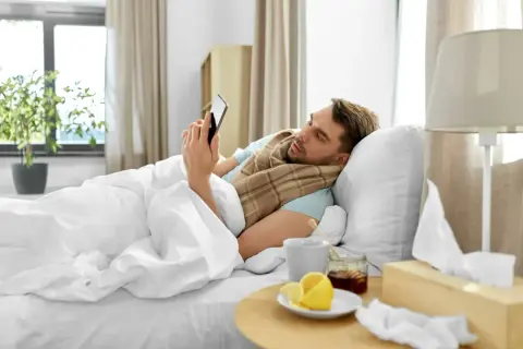 Chory mężczyzna w łóżku, owinięty kocem, przegląda telefon. Obok kubek z herbatą, cytryna i miód. To zwolnienie siła wyższa.