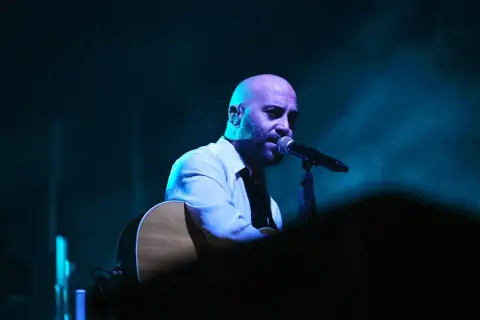 Giuliano Sangiorgi dei Negramaro, con la sua chitarra, canta al microfono, un passo indietro sul palco illuminato da luci blu.