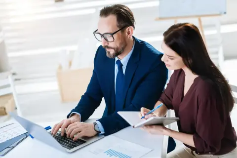 Biznesmeni pracują nad inwentaryzacją, mężczyzna pisze na laptopie, kobieta notuje.