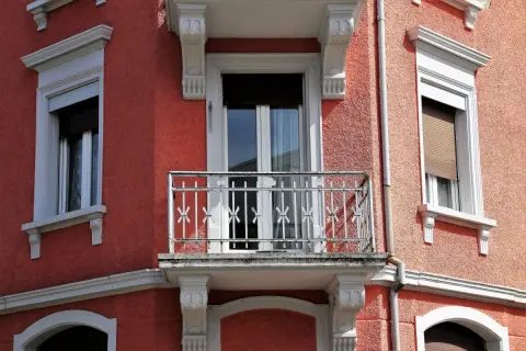 Dettaglio di un muro esterno con finitura a rasatura, finestre con persiane e un piccolo balcone con ringhiera in ferro battuto.