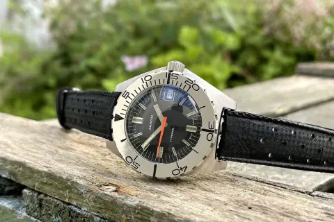 Reloj Sandoz Geneve Typhoon con correa negra, sobre madera rústica y fondo verde.