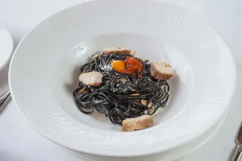 Pasta negra con trozos de pescado, huevas de salmón y yema de huevo. La tinta de calamar, ingrediente principal, aporta un sabor único y propiedades beneficiosas.