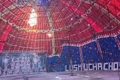 Interior de carpa de circo con techo rojo y azul, telón de fondo de acróbatas y luces que forman "MUCHACHOS". ¡Un espectáculo en Bempota, Ourense!