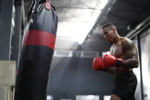 A boxer training intensely, sweat glistening on his skin as he throws punches at a heavy bag in a dimly lit gym.