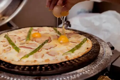 Ręka wylewa jajko na pizzę z boczkiem i szparagami. Pyszna pizza z jajkiem w trakcie przygotowania.