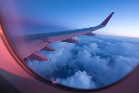 View from an airplane window at sunset. The wing stretches across the frame, bathed in pink and purple light, over a sea of clouds.